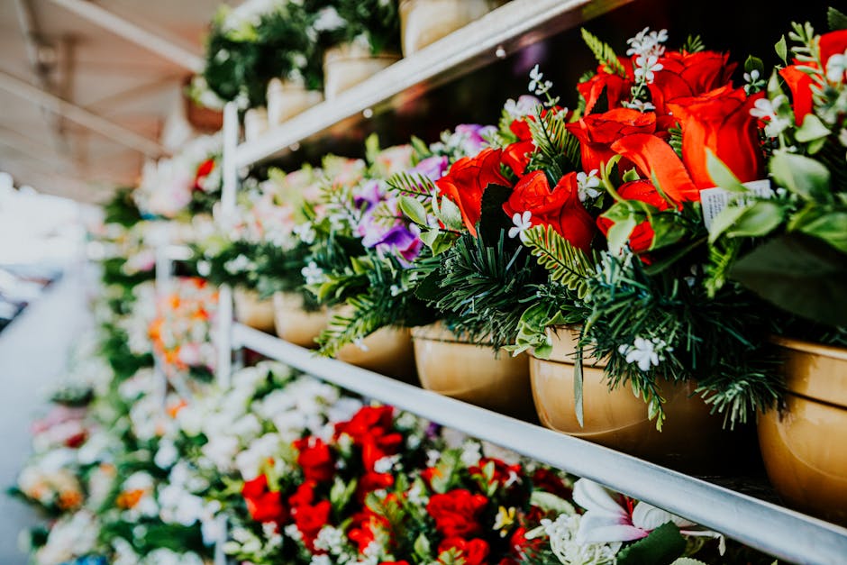 A detailed floral display featuring vibrant red roses, white and purple flowers, and assorted green foliage arranged in beige ceramic pots on a metal shelf at a flower shop. The bouquet showcases fres