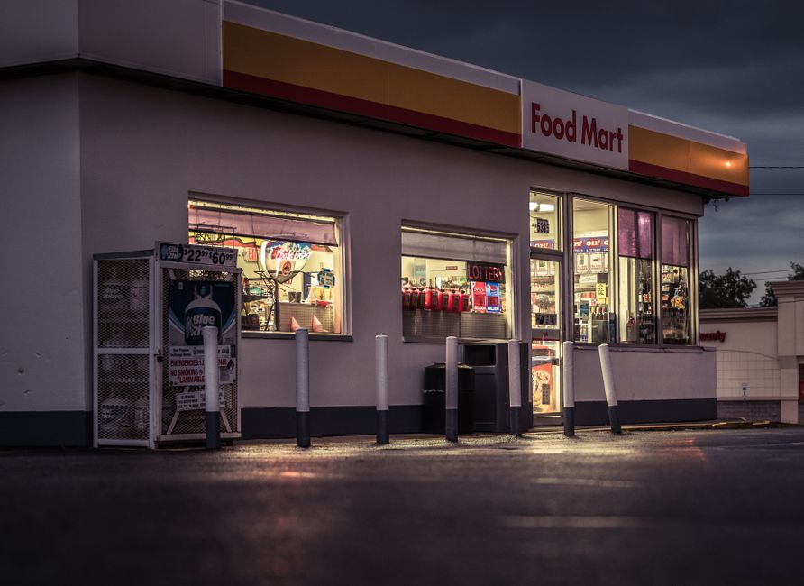 A close-up view of the exterior of a convenience store with a sign reading 'Food Mart' on a dark, cloudy evening. The store has large glass windows displaying brightly lit interior shelves stocked wit
