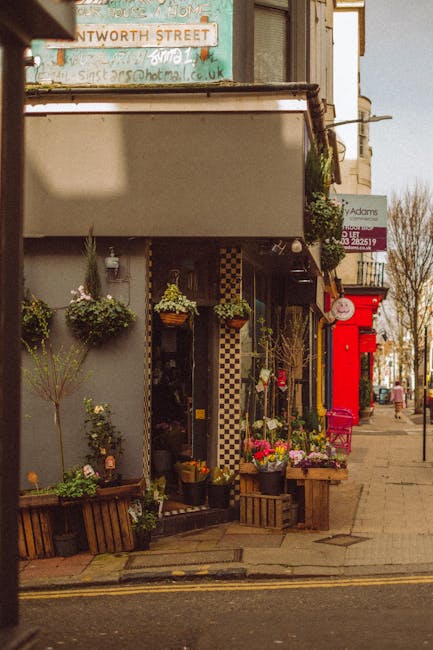 The exterior of a flower shop on a city street features an inviting display of vibrant floral arrangements and potted plants arranged on wooden crates and hanging baskets. The flowers include bright p