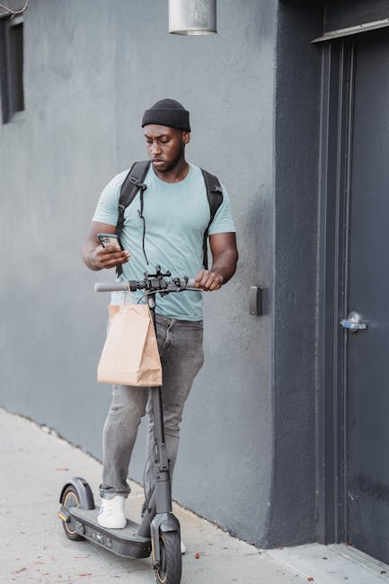 A young man with dark skin, a black beanie, and a light blue T-shirt stands outdoors next to a gray building wall. He holds a smartphone in his right hand and a small paper bag in his left. He is stan
