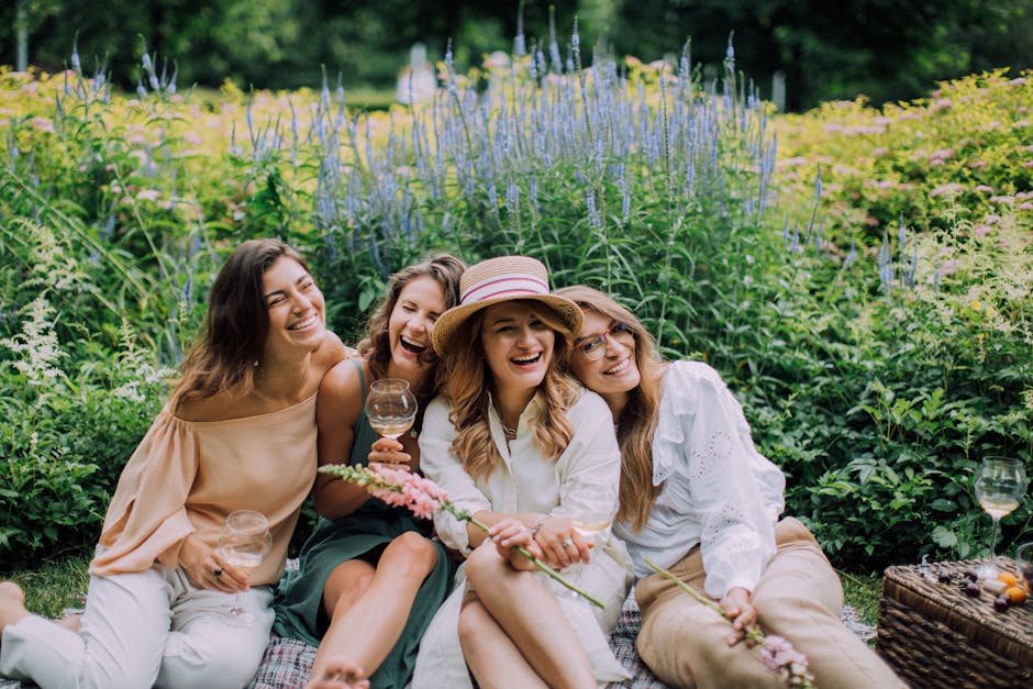 Four women seated on a picnic blanket outdoors surrounded by lush green foliage and flowering plants, enjoying a cheerful moment together. They are dressed in light, summery clothing; one wears a beig