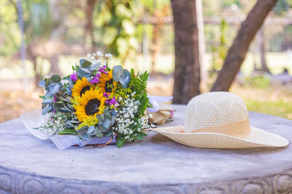 A vibrant floral bouquet featuring sunflowers, purple statice, white gypsophila, and eucalyptus leaves arranged in a wrapping paper with a white ribbon, placed on a weathered wooden table outdoors. Ne